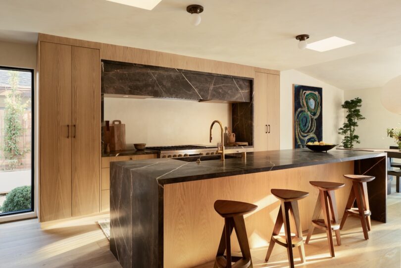 Modern kitchen with a large island featuring a dark marble countertop, wooden barstools, and light wood cabinetry. A painting and greenery are in the background, under natural light.