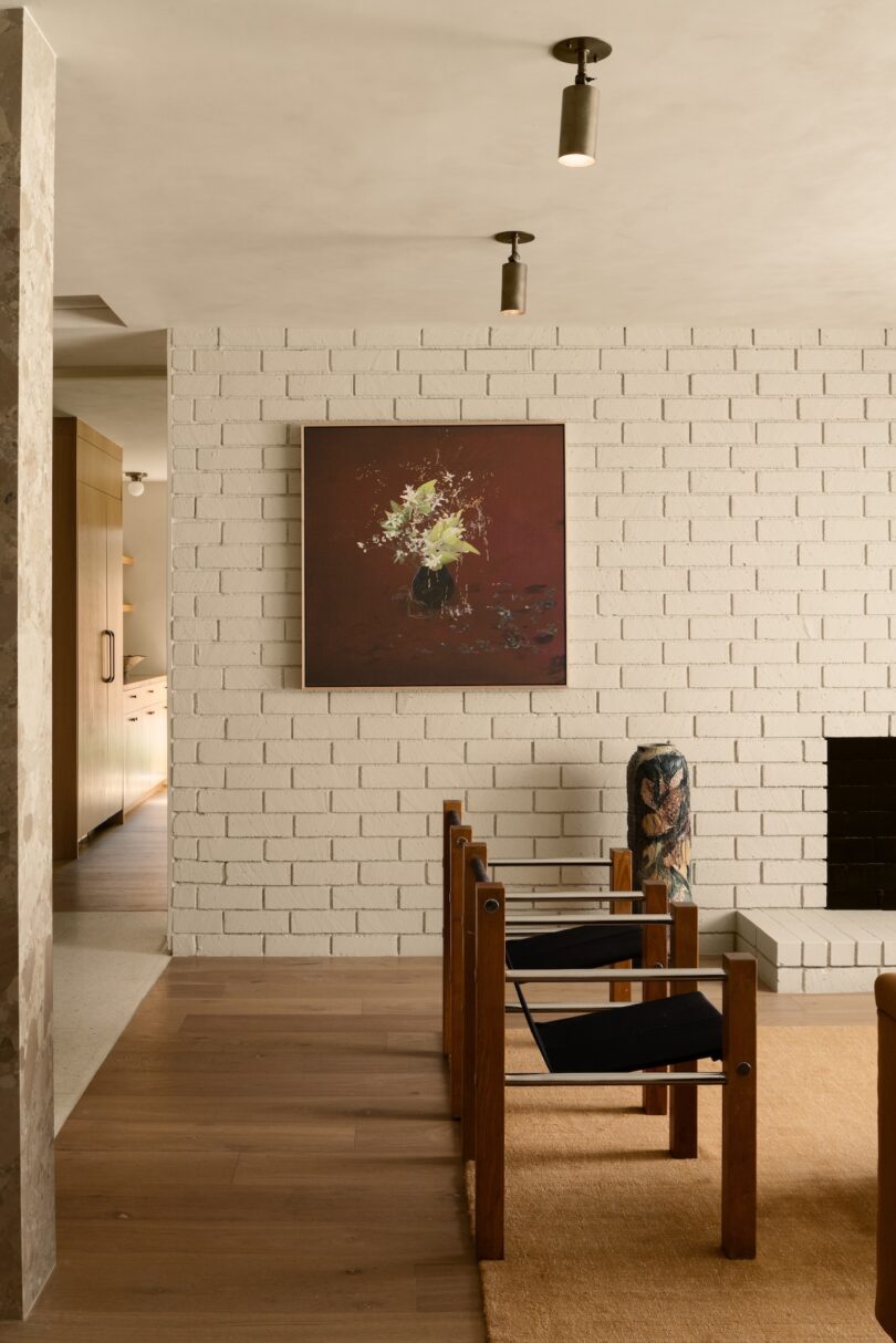 Interior with white brick wall, abstract painting, two wooden-framed chairs, and a dark, cylindrical vase.