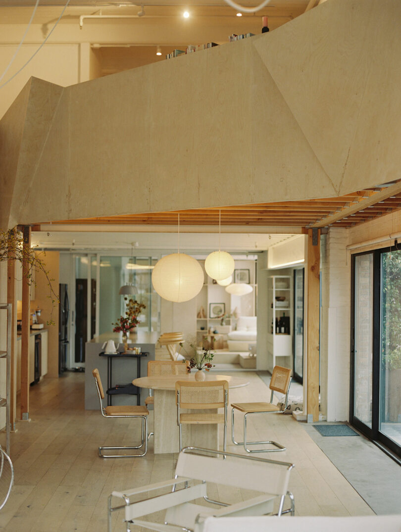 Modern open-concept living space designed by Amanda Gunawan, featuring wooden accents, round paper lanterns, a dining table, and stylish chairs. Glass doors seamlessly connect to an inviting outdoor area.