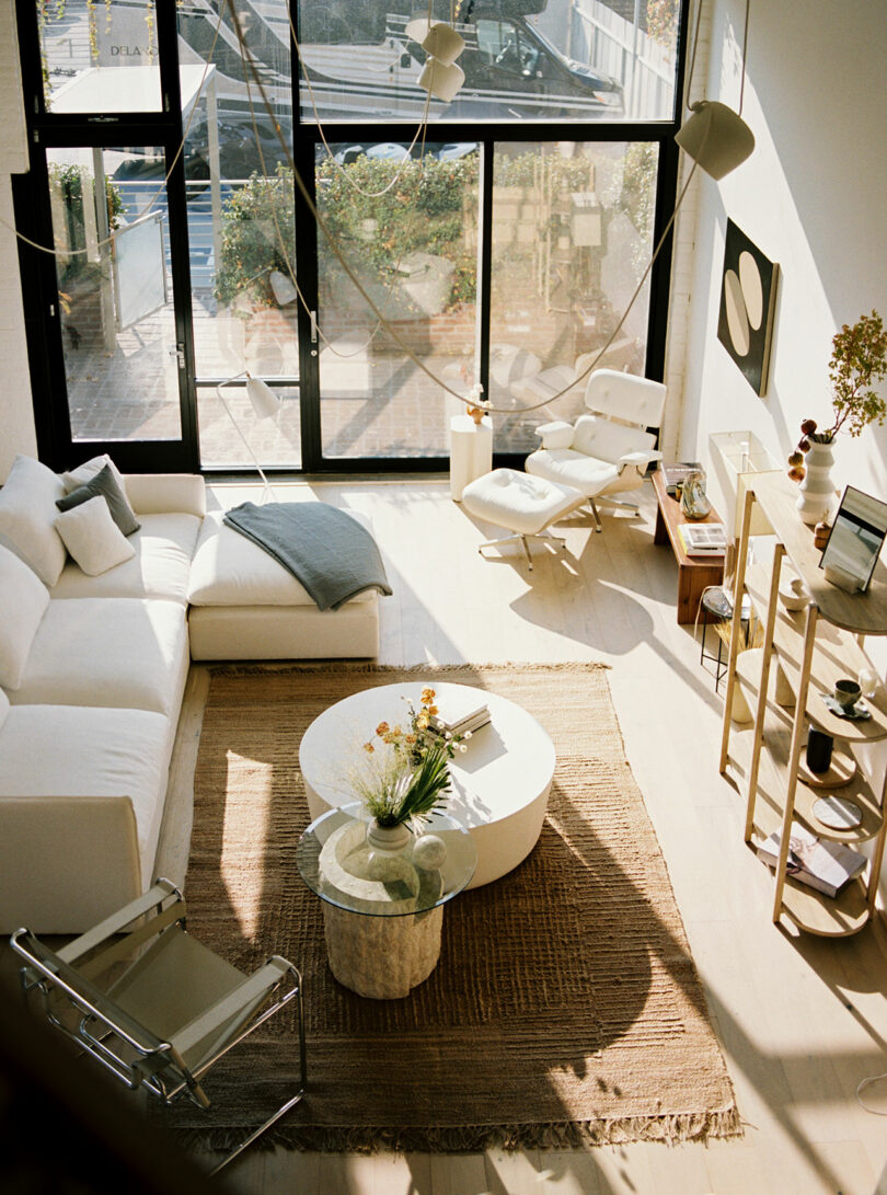 Designed by Amanda Gunawan, the bright living room features large windows, a white sectional sofa, and a round coffee table atop a woven rug. Chairs and carefully selected decor items accent the space as sunlight streams through glass doors.