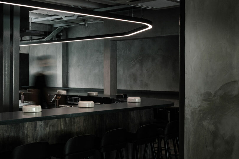 Dimly lit modern bar designed by Amanda Gunawan, featuring sleek black furniture and a curved overhead light fixture. A blurred figure is seen in motion behind the bar, adding dynamic energy to the sophisticated ambiance.