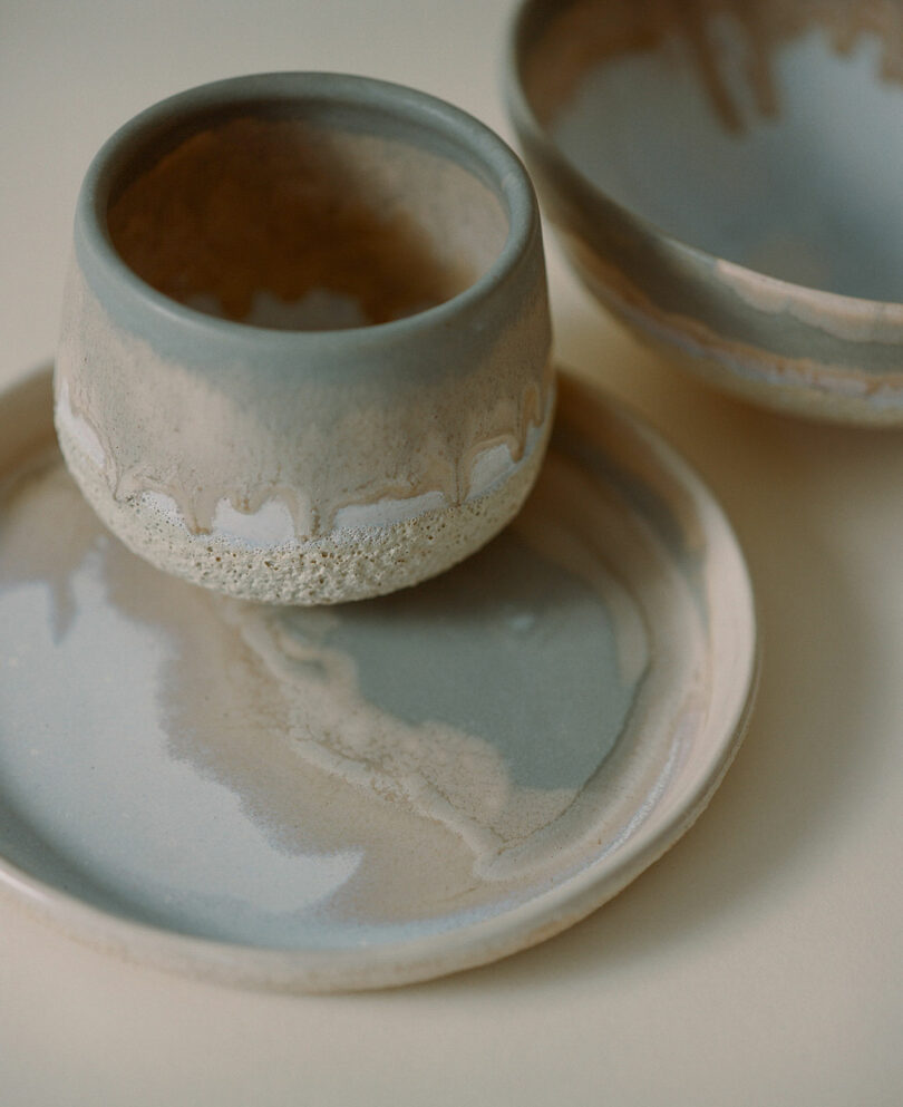 Ceramic dishes with a speckled, earthy glaze, including a small cup, bowl, and plate by Amanda Gunawan, displayed on a light surface.