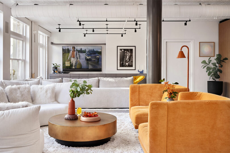 Modern living room with white and orange seating, round coffee table with decor, large windows, wall art, and plants.