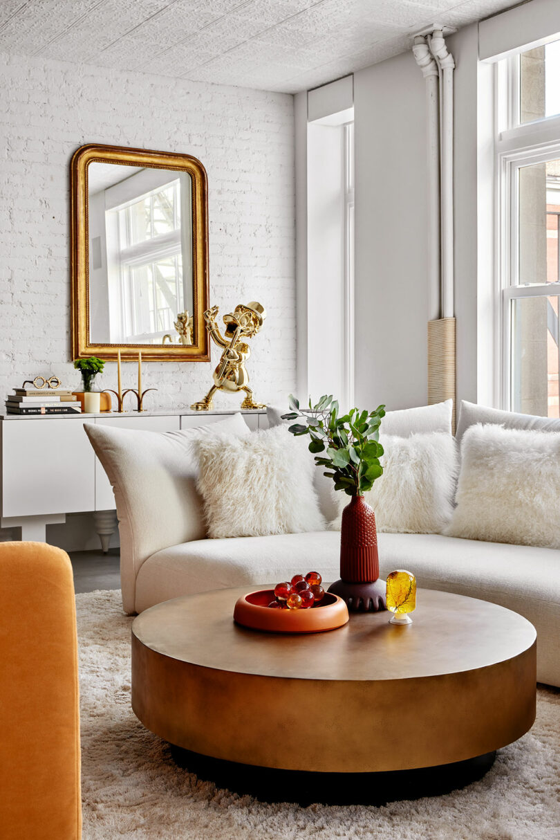 Modern living room with white couch, fluffy pillows, and a round gold coffee table. Decorative items include a gold sculpture, a vase with greenery, and a large mirror on a white brick wall.