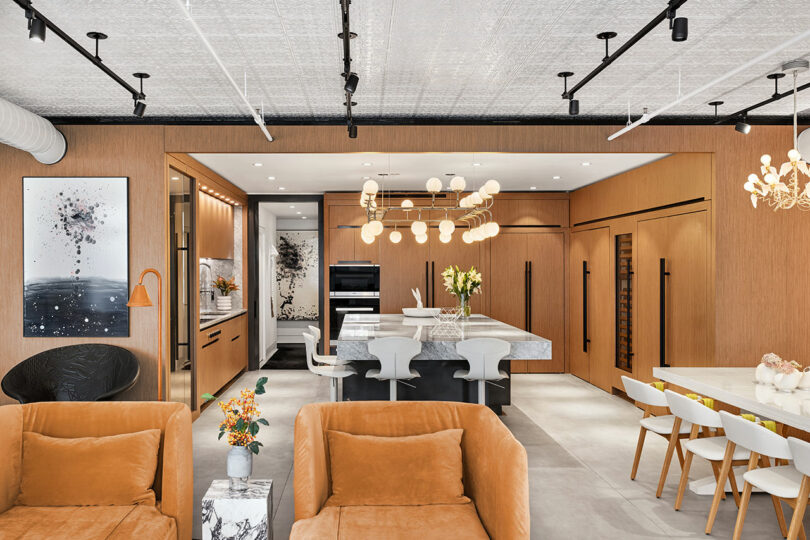 Modern kitchen and living area with wooden cabinets, marble countertops, pendant lighting, and tan sofas. Dining table with white chairs and decorative flowers on tables.