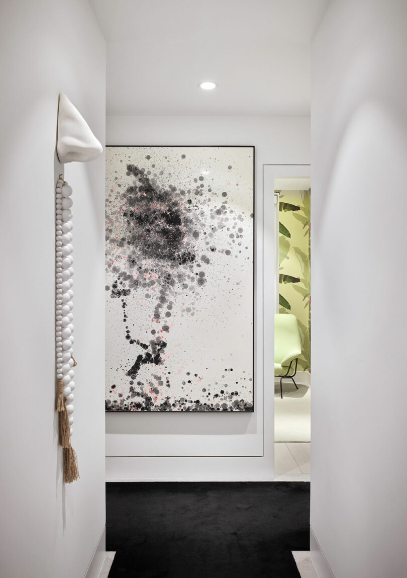 A hallway with a large abstract painting featuring black and pink dots. A wall hanging with white beads and a tassel is on the left. An open door reveals a room with a green chair.