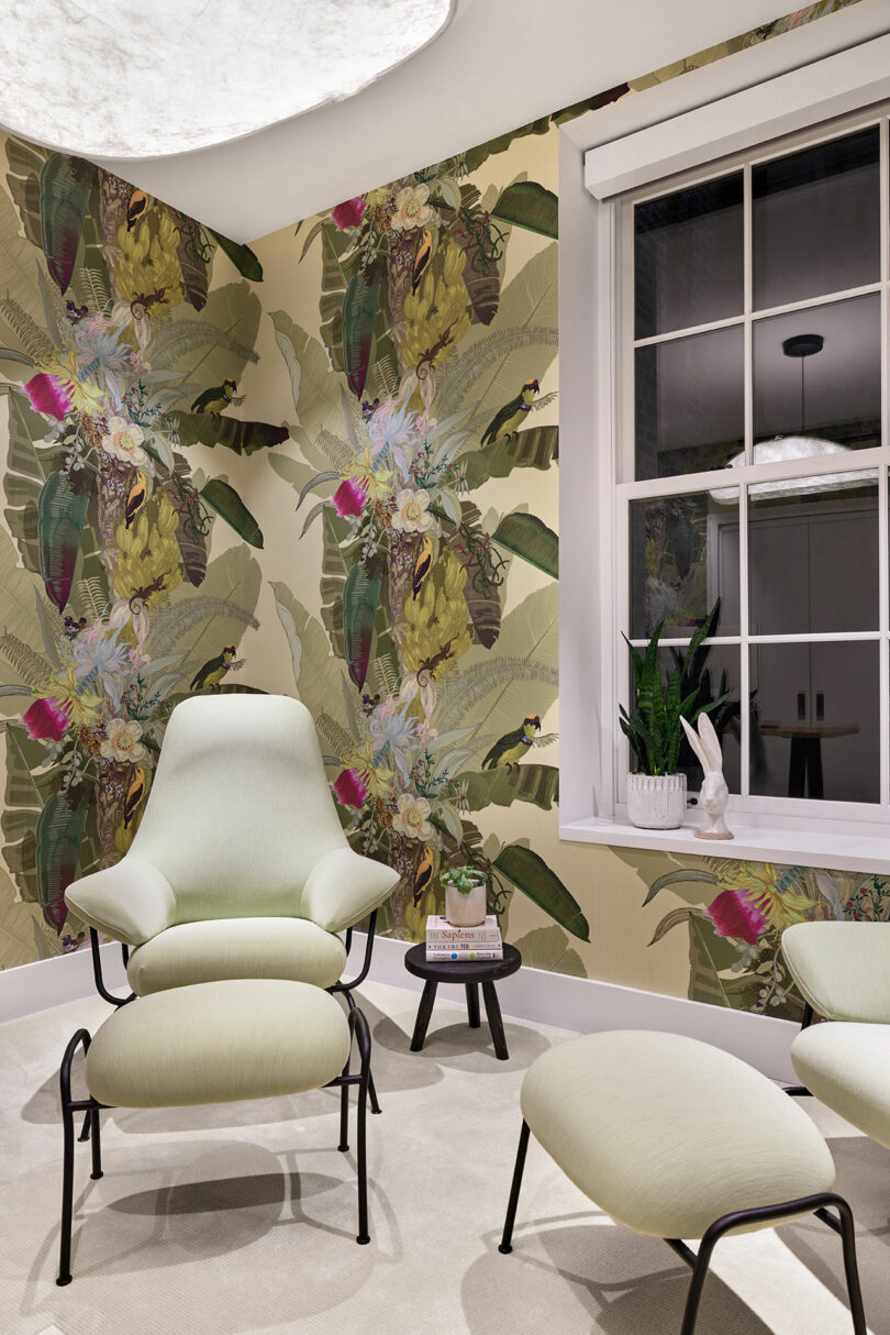 A room with botanical wallpaper, featuring a green armchair, matching footstool, small round table with a plant, and a window.