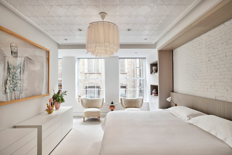 A modern bedroom with a white bed, textured ceiling, large windows, two armchairs, a dresser with decorative items, and a framed artwork on the wall.