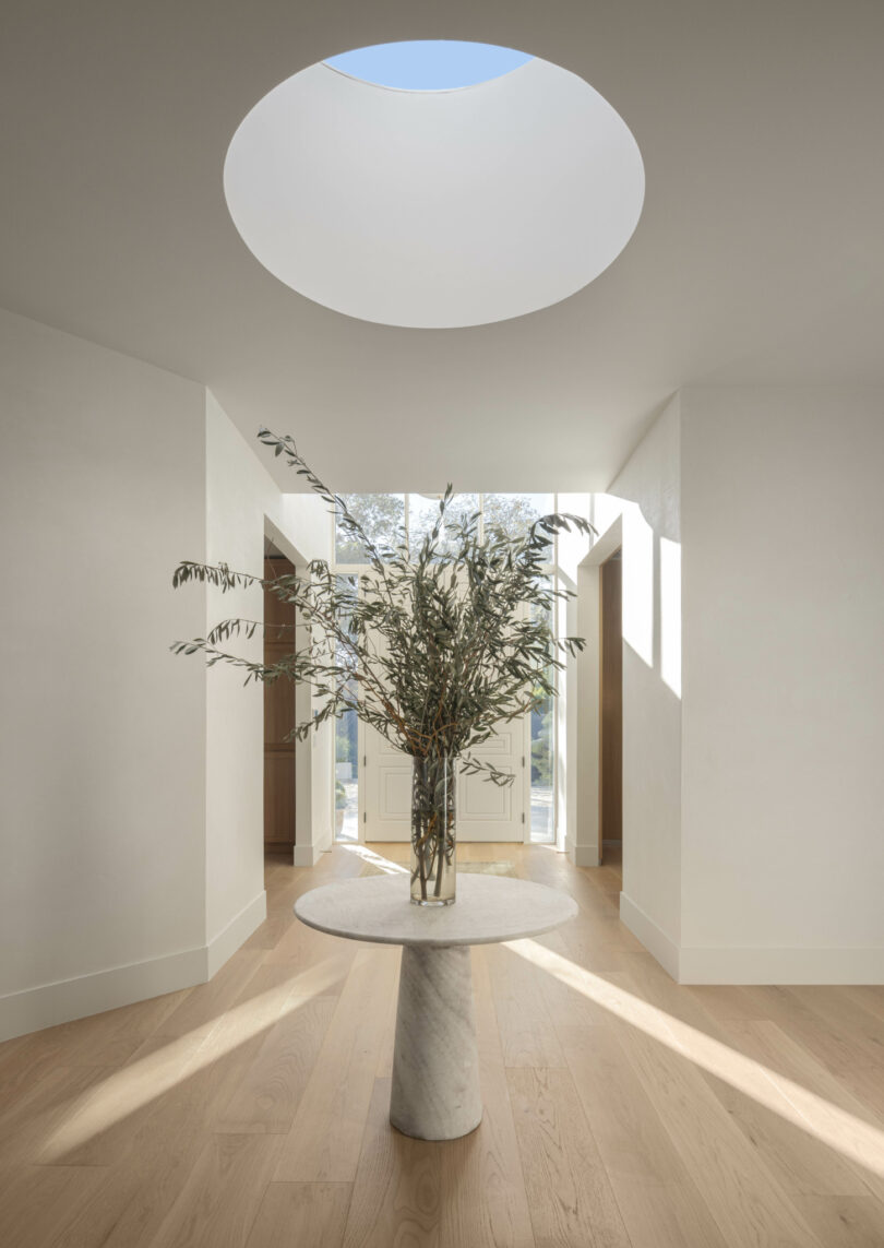 A minimalist hallway by Studio OSKLO features a round skylight, a column with a vase of greenery, light wood floors, white walls, and a door with sidelights in the background.