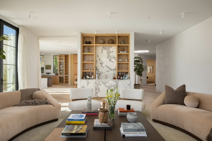 A modern living room featuring curved beige sofas, a central coffee table with books and decor, and a wooden shelving unit with decorative items against a marble backdrop designed by Studio OSKLO.