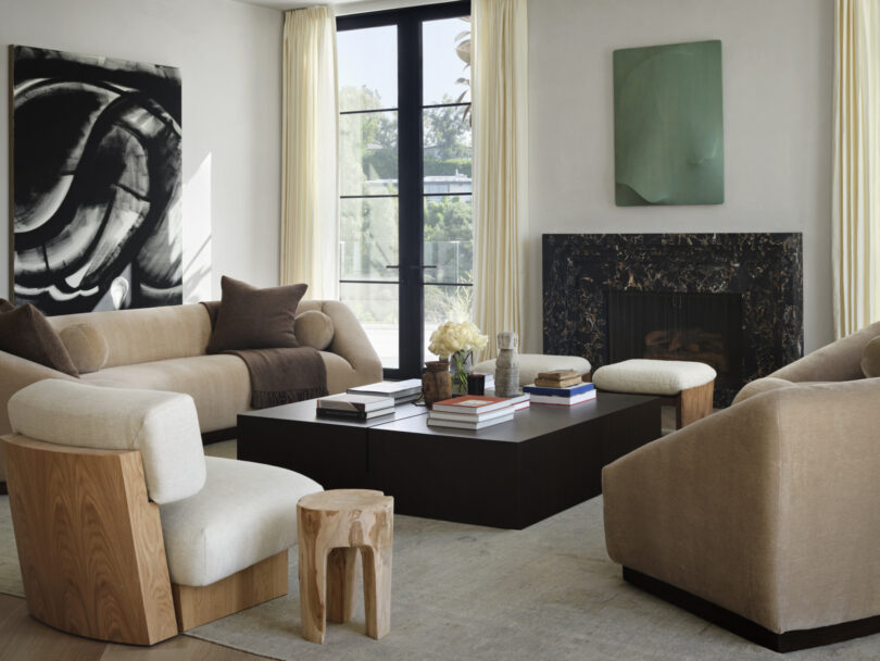 A modern living room designed by Studio OSKLO features beige sofas, a black square coffee table, abstract paintings, large windows with light curtains, and a black marble fireplace.