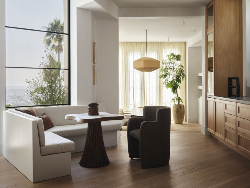 A modern, sunlit dining area designed by Studio OSKLO features a corner bench, two chairs, a round table, large windows with city views, wooden cabinets, and indoor plants.