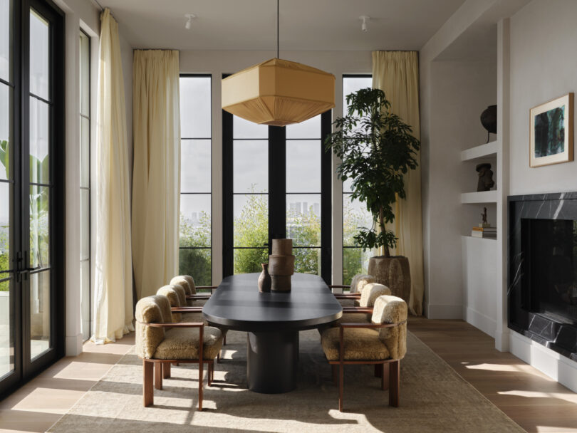 A modern dining room designed by Studio OSKLO features a large black table, six beige chairs, tall windows, a geometric light fixture, and a large potted plant. Shelving and a fireplace adorn the right side of the room.