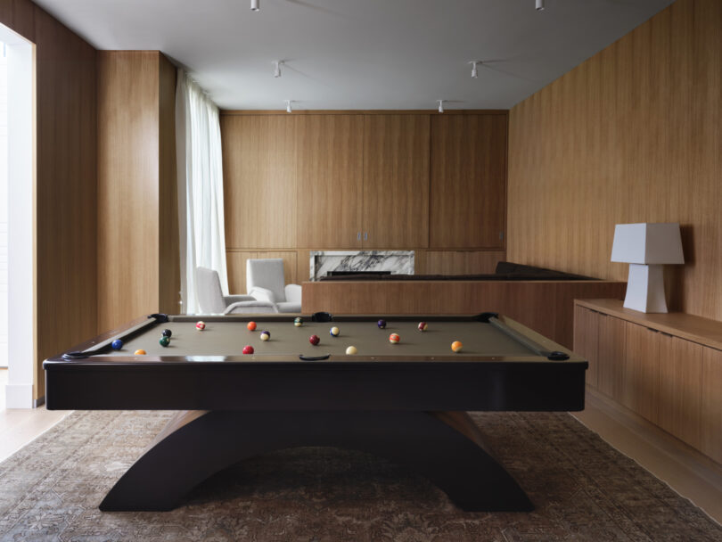 A modern room designed by Studio OSKLO features wood-paneled walls, a pool table, a marble fireplace, a few white chairs, and a lamp on a cabinet.