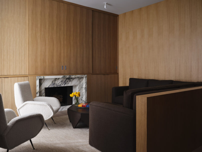 Modern living room designed by Studio OSKLO, featuring brown wood-paneled walls, white armchairs, dark brown sofas, a marble fireplace, and a black coffee table adorned with a stack of books and a vase of yellow flowers.