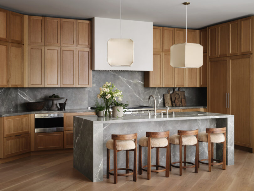 A modern kitchen by Studio OSKLO features wooden cabinets, a marble island with four upholstered stools, pendant lights, and built-in appliances against a backdrop of stone backsplash and countertops.