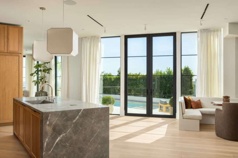 Modern kitchen with an island featuring a marble countertop, large glass doors opening to a patio, and a seating area with a couch and chair, all flooded with natural light. The design embodies the sleek elegance synonymous with Studio OSKLO.