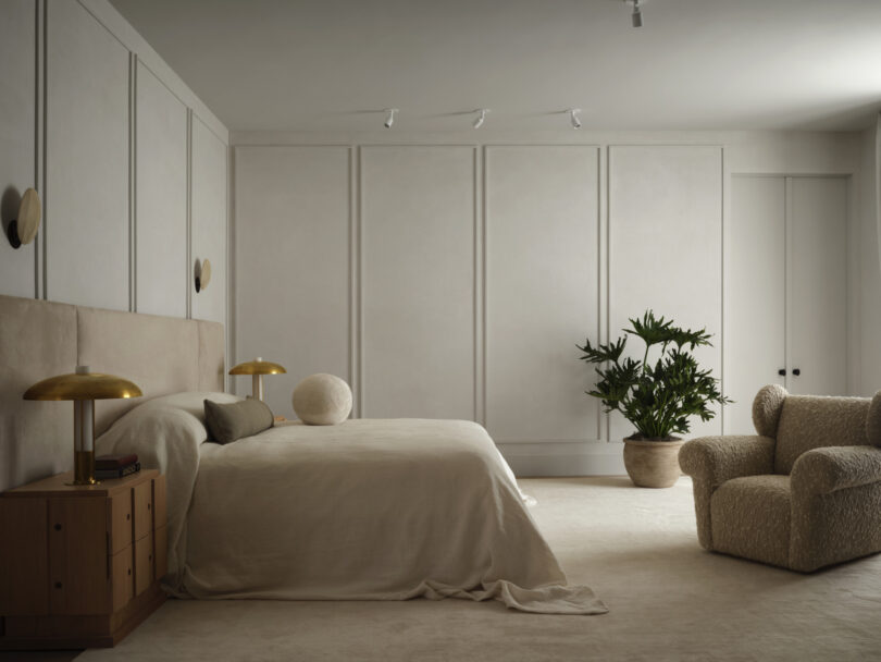 A minimalist bedroom with neutral tones by Studio OSKLO features a bed with beige linens, a nightstand with gold lamps, an armchair, a large potted plant, and wall paneling.