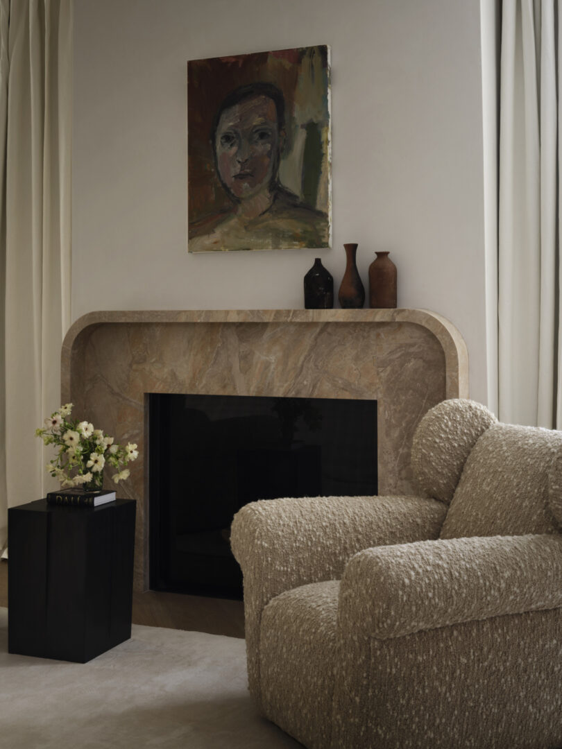 A beige textured armchair by Studio OSKLO sits beside a black side table with flowers, in front of a marble fireplace with vases and a painted portrait hanging above it. Cream curtains frame the setting.