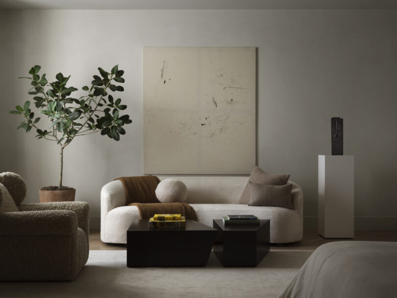 A minimalist living room by Studio OSKLO features a white sofa, abstract wall art, a tall potted plant, two black coffee tables, a beige armchair, and a sculpture on a pedestal.