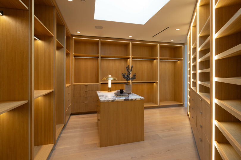 A spacious walk-in closet by Studio OSKLO features wooden shelving and drawers, an island countertop with a lamp and décor, and a skylight providing natural light.