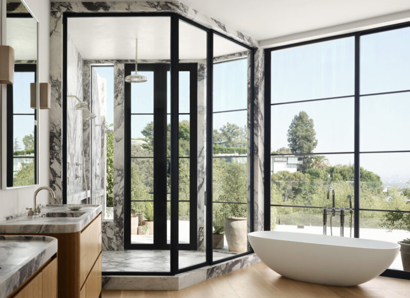 Modern bathroom by Studio OSKLO featuring a freestanding bathtub, double vanity with wood cabinets, and a glass-enclosed shower. Large windows provide natural light and a view of greenery outside.