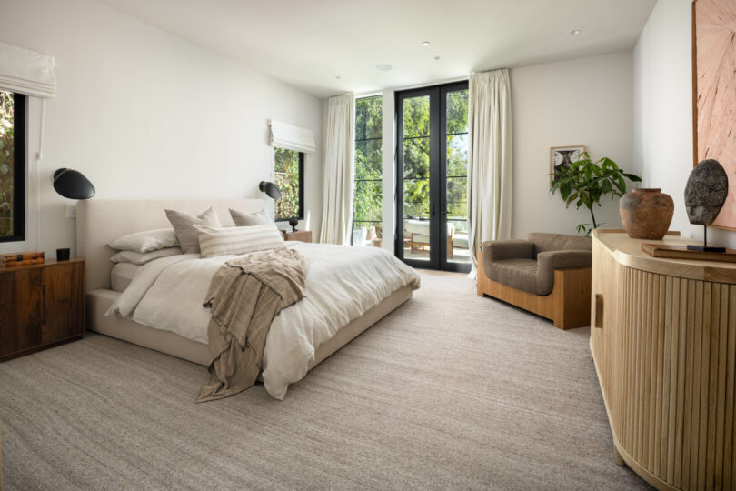 A modern bedroom designed by Studio OSKLO features a large bed with neutral-colored linens, a cushioned chair, wooden furniture, a potted plant, and floor-to-ceiling windows offering a tranquil view of trees outside.