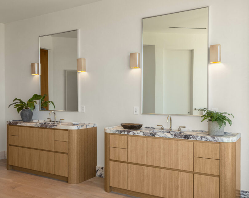 A modern bathroom with two wooden vanities, designed by Studio OSKLO, each featuring a rectangular mirror and a wall sconce above. Countertops display marble patterns and are adorned with small plants for decor, complemented by light-colored walls and flooring.