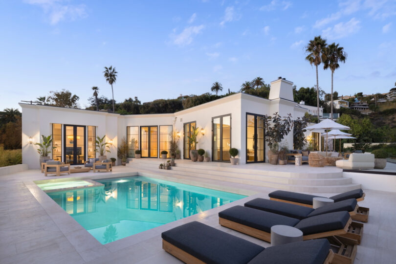 Modern white villa with large windows, designed by Studio OSKLO, surrounded by palm trees and featuring a spacious outdoor area with a swimming pool, lounge chairs, and patio furniture under umbrellas.