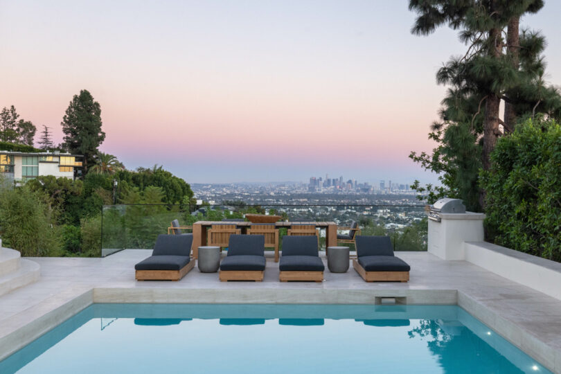 A modern poolside area by Studio OSKLO features four lounge chairs, a wooden dining set, and views of a distant city skyline at dusk. Tall trees and lush greenery surround the space.