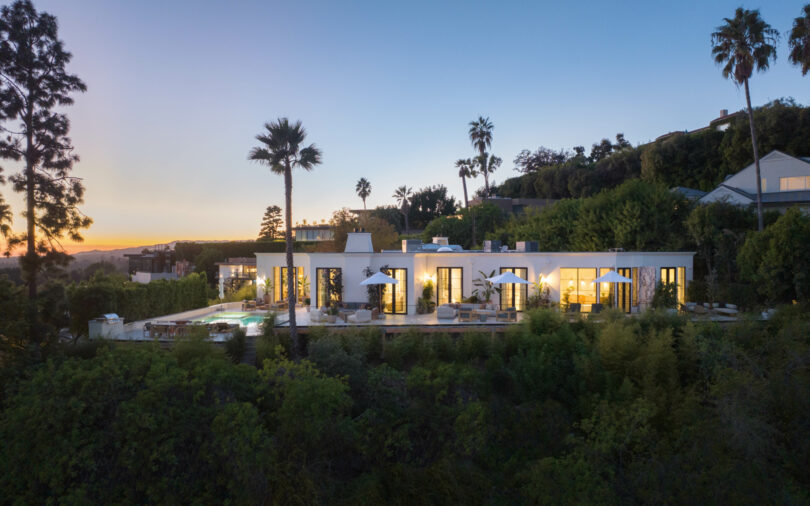 Spacious modern hillside mansion designed by Studio OSKLO, featuring large windows, palm trees, and an outdoor patio, all illuminated under an evening sky.