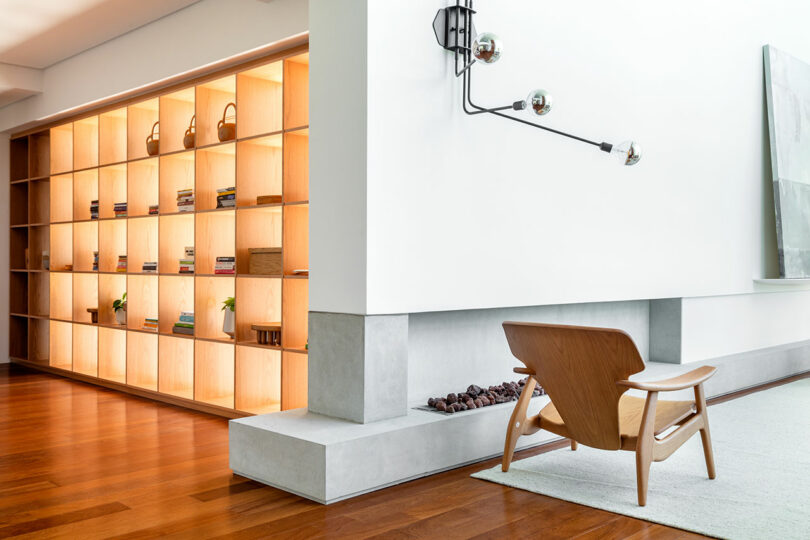 Modern living room with wooden floors, a minimalist wood and fabric chair, built-in shelving unit with illuminated compartments, and a white wall featuring a contemporary light fixture.