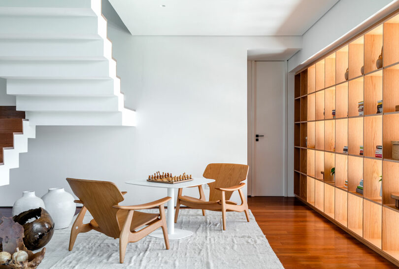 A modern room with two wooden chairs and a round white table with a chessboard on it. A wooden bookshelf with books lines one wall, and a white staircase is adjacent. Floor is wooden with a light rug.