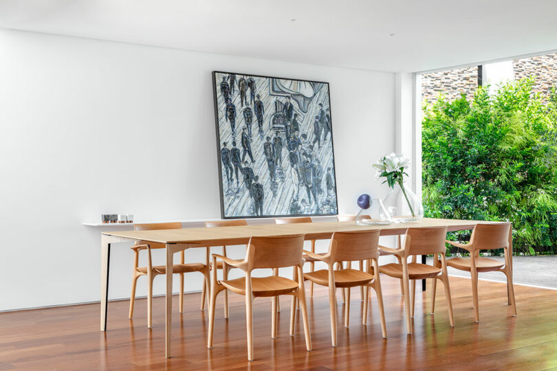 Modern dining room with a long wooden table, six wooden chairs, large abstract painting on the wall, and a vase with white flowers. A large window with lush greenery outside provides natural light.