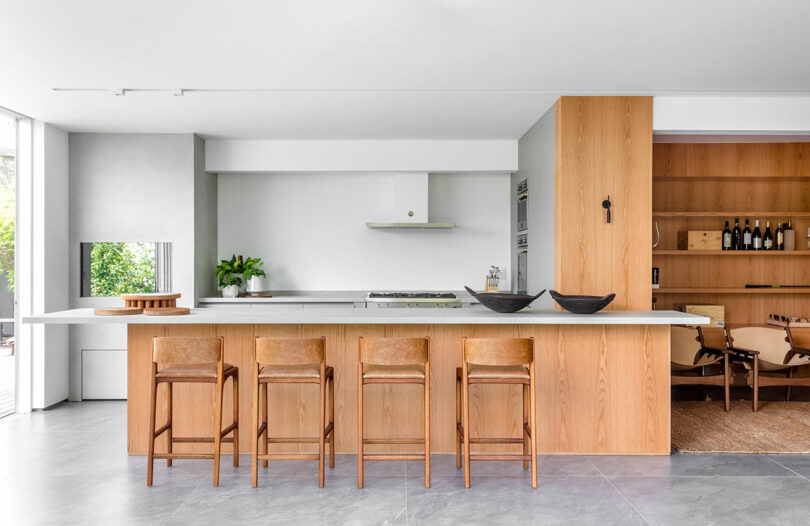 Modern kitchen with a large island, wooden cabinets, and four wooden barstools. There is a shelf with bottles to the right and greenery visible outside through a window on the left.