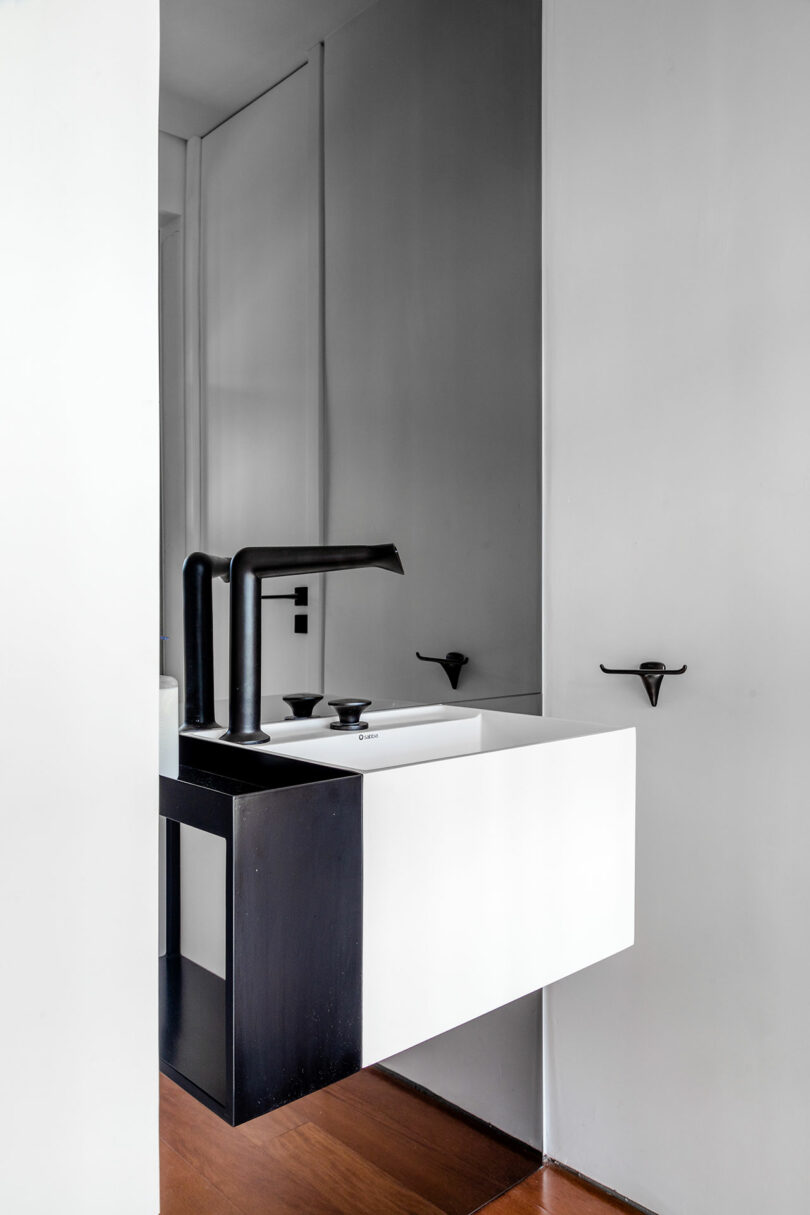 Modern minimalist bathroom with a black and white floating sink, black faucet, and mirror above. White walls and wood floor present a clean, sleek aesthetic. Wall-mounted fixtures are visible.