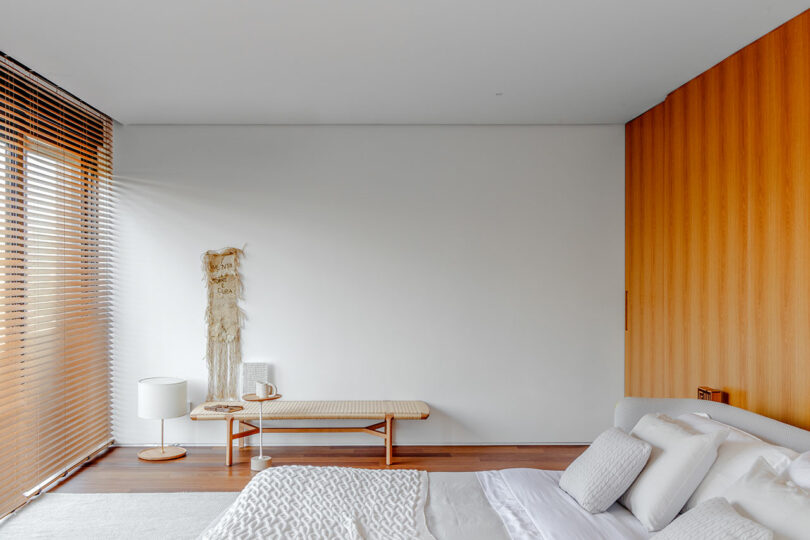 A minimalist bedroom with a white bed, light wood bench, bedside table with a lamp, wooden blinds, and a woven decor piece on a white wall. The room has a clean and simple design.