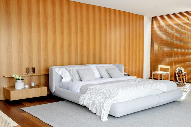 A minimalist bedroom with a low-profile bed, white bedding, wooden nightstand, striped wooden wall paneling, a gray rug, and window blinds with natural light filtering in.