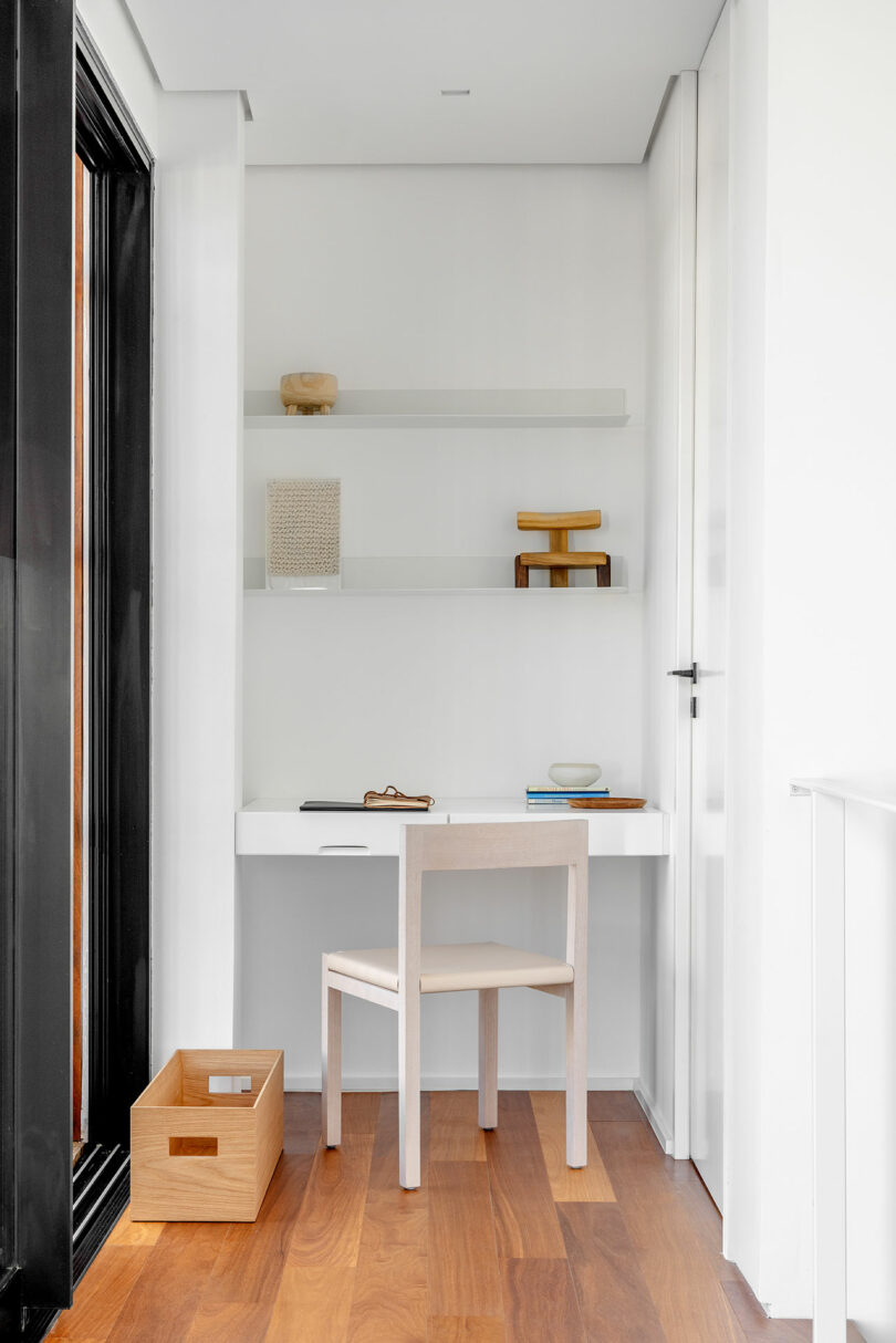 A minimalist workstation with a white desk and chair, flanked by floating shelves holding decorative items. The space features wood flooring and a window on the left. A wooden box sits on the floor.