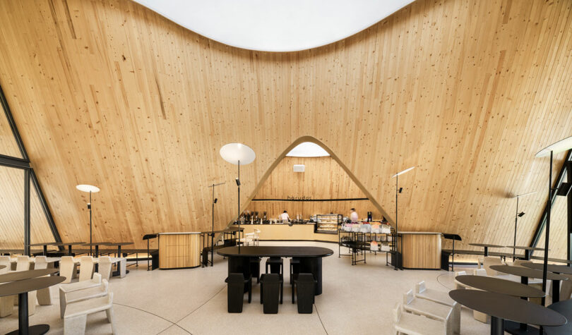 A modern, wooden interior cafe with a high, curved ceiling, central round table, various seating options, and a visible counter where two people are working.