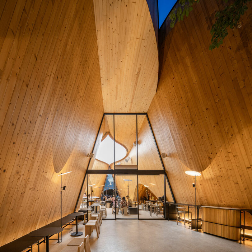 Interior of a modern building with wooden triangular walls, large glass entrance, and minimalist furnishings under warm lighting.