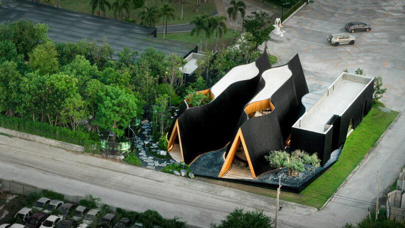 Aerial view of a modern black-roofed building with unique wave-like designs, surrounded by greenery and adjacent to a parking lot with a few cars.