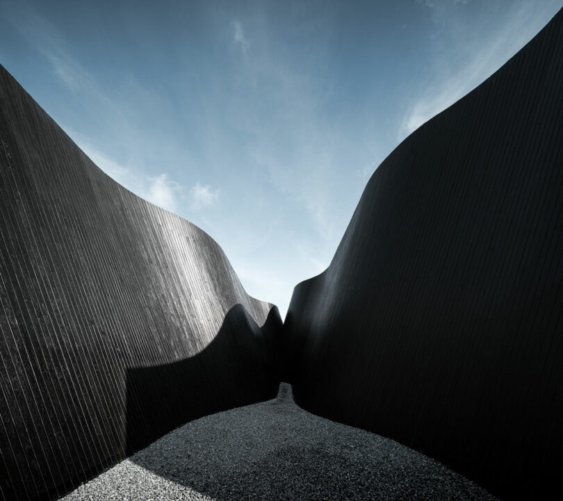 Curved black wall structures rise against a clear blue sky, creating a narrow pathway covered in gravel.