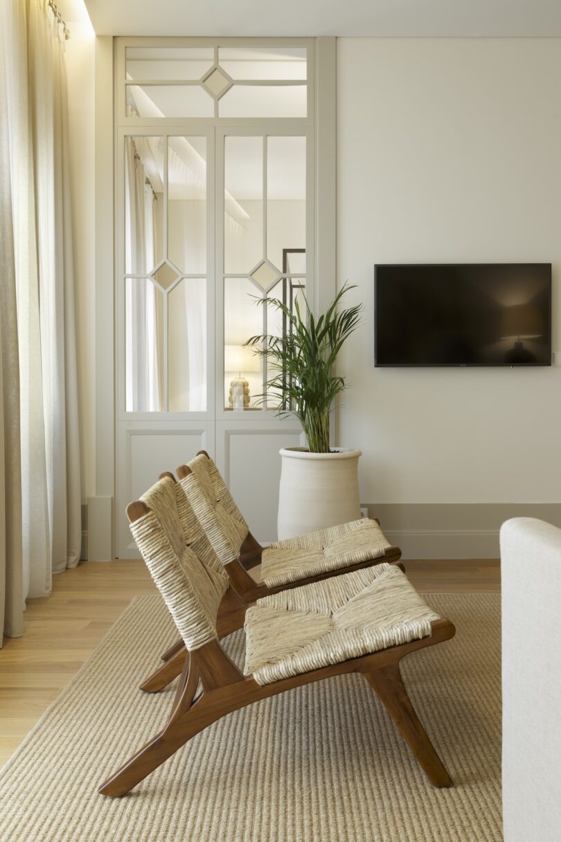 Seating area with two woven chairs, a potted plant, and a wall-mounted television, framed by large windows with sheer curtains