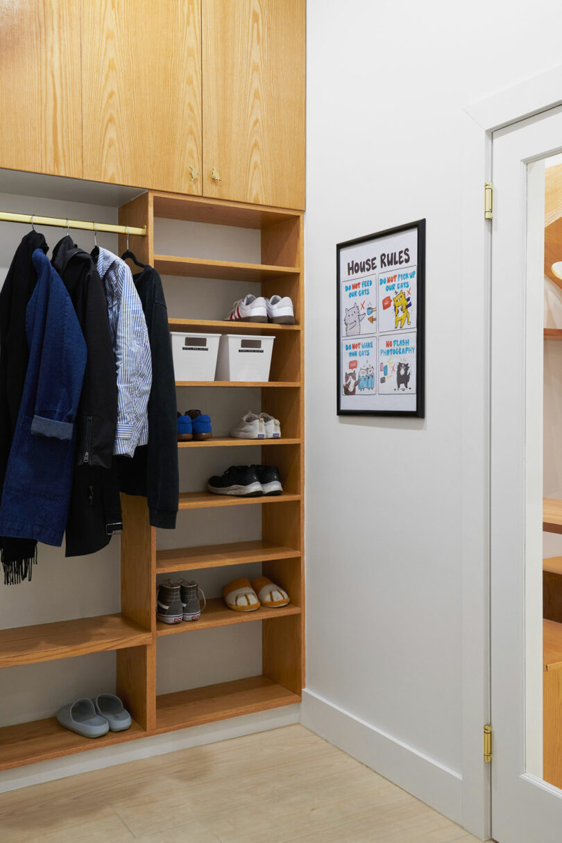 Entryway with wooden shelving for shoes and coats, featuring jackets, shirts, and various footwear. A framed "House Rules" sign is displayed on an adjacent wall.