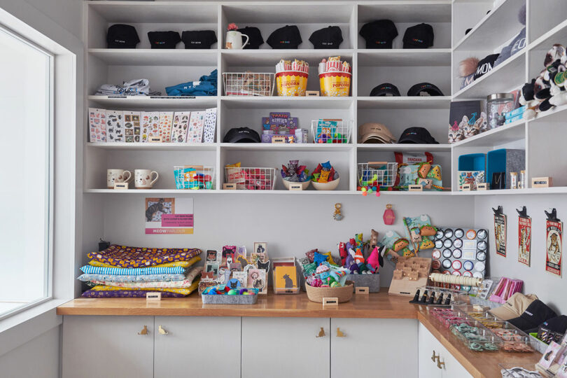 A store display shelf filled with colorful items including hats, mugs, fabrics, toys, and stationery, organized in baskets and on surfaces. Brightly lit with natural light from a window on the left.