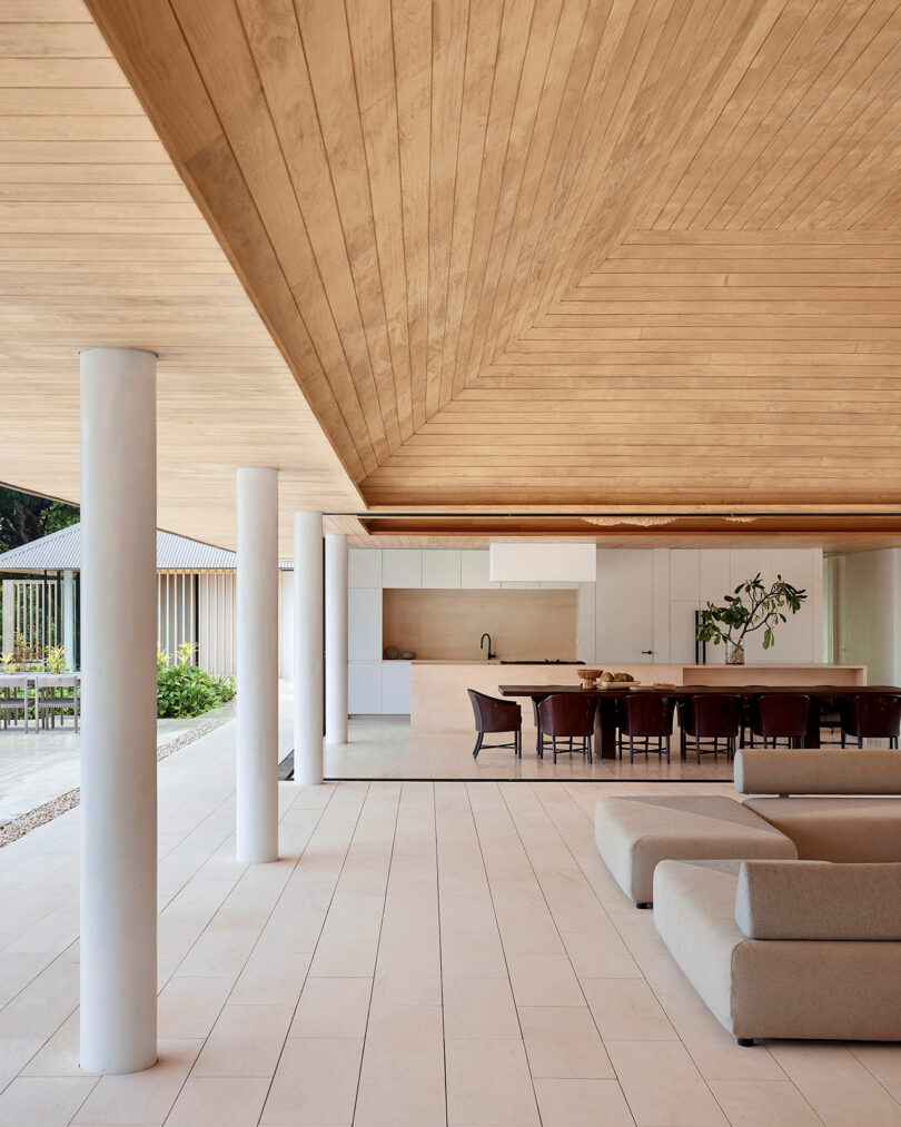 Spacious modern interior featuring a wooden ceiling, white pillars, tile flooring, a gray sofa, a dining table with several chairs, and a kitchen area with white cabinets and countertops.