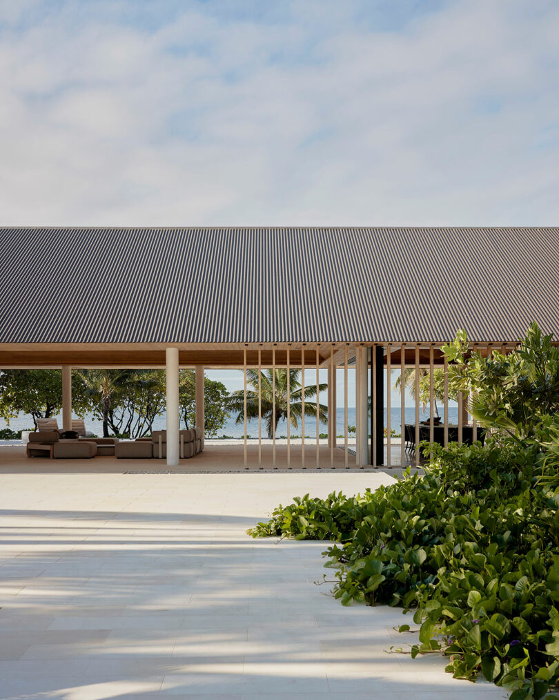 Modern open-air building with a slanted roof, featuring outdoor seating areas. Green foliage in the foreground and a view of the ocean and trees in the background.