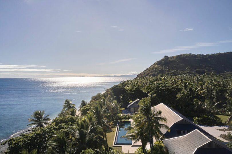 A coastal resort with buildings surrounded by palm trees overlooks a calm, sunlit ocean and a mountainous area on the horizon.