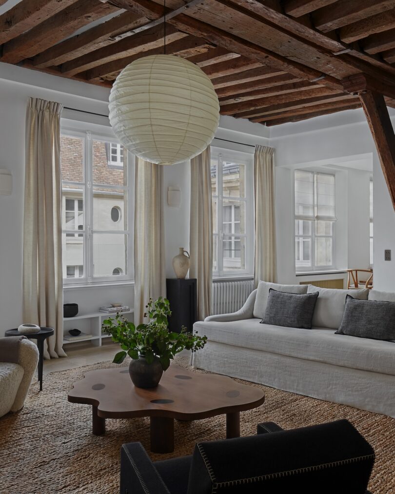 A bright and airy living room with exposed wooden beams, linen curtains, and a cozy seating area